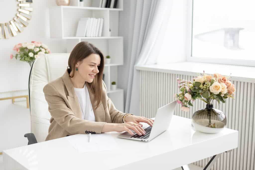 a young female coach works in a home office in a light interior with a laptop and flowers