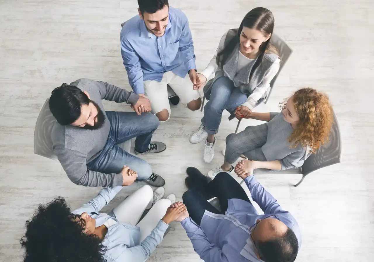 Top-down view of a diverse group of people holding hands in a circle during a group therapy session, symbolizing connection, support, and healing.